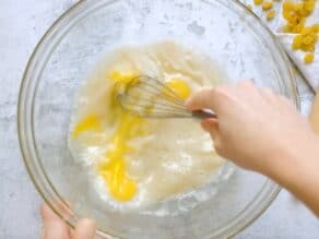 Hand whisking together eggs, water, honey and yeast in glass bowl on marble countertop.