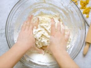 Hands kneading loose dough with flour in glass bowl on marble countertop.