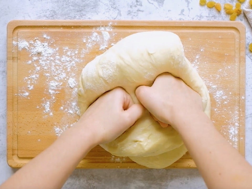 Hands kneading puffy challah dough on floured wooden board on marble countertop.