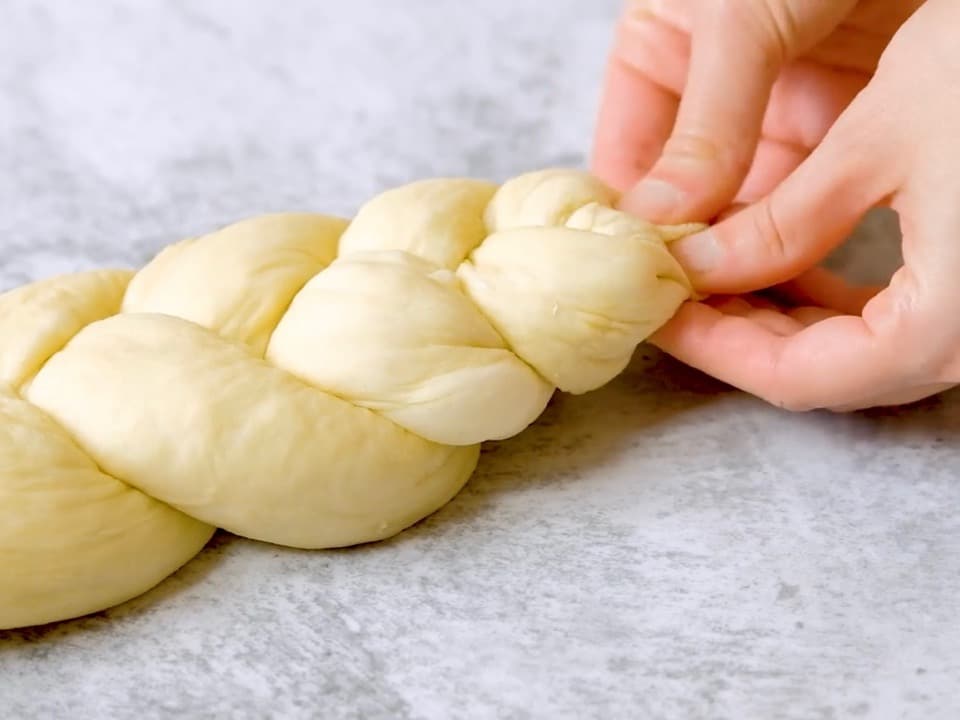 Two hands braiding the ends of a three strand challah dough braid.