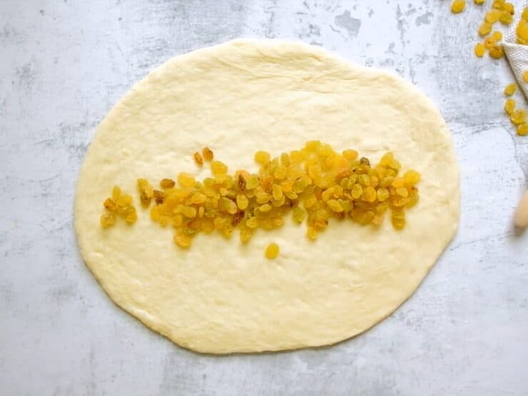 Overhead shot of challah dough that has been rolled into an oval shape. Golden raisins are spread horizontally across the center of the dough.