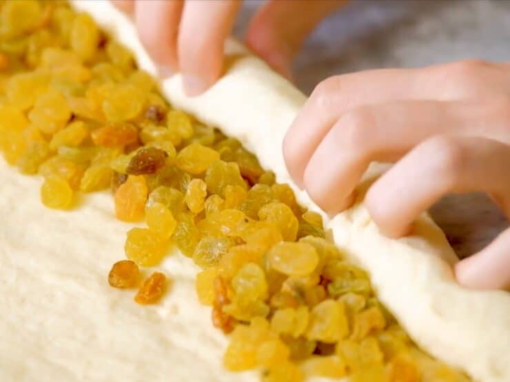Two hands rolling challah dough filled with raisins into a single strand.