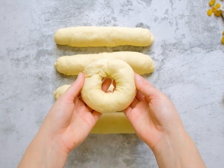 Two hands forming a ring-shaped circle of challah dough, four short thick dough strands on counter in background.