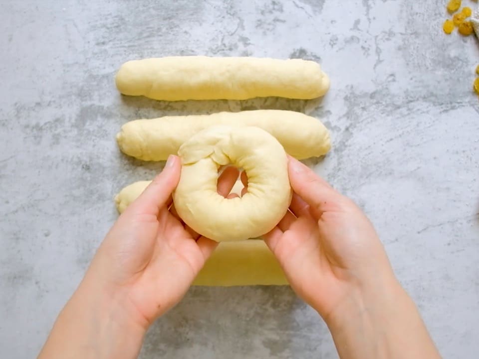 Two hands forming a ring-shaped circle of challah dough, four short thick dough strands on counter in background.