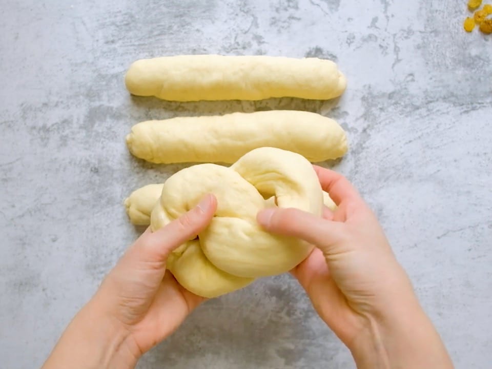 Two hands connecting two loops of challah dough, three short thick dough strands on counter in background.