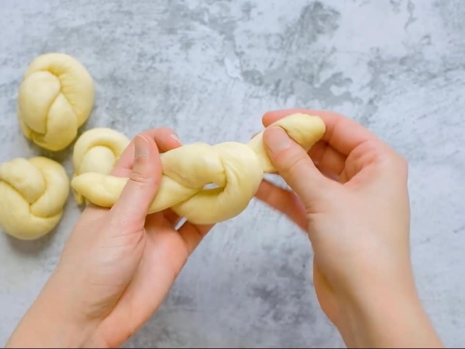 Hand pulling small strand of dough into the first stage of a knot to create a roll shape; three challah knots unbaked on countertop in background.