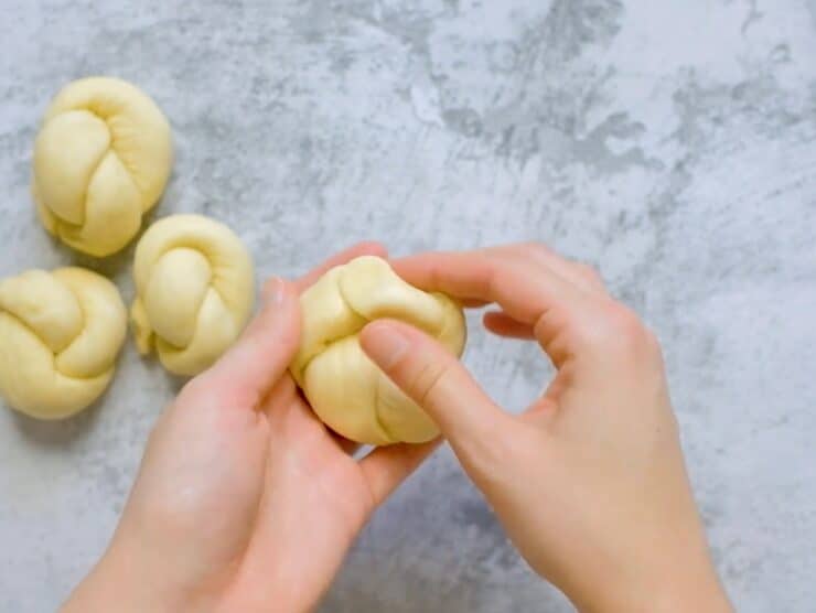 Hand closing a knot of dough to create a roll shape, three challah knots unbaked on countertop in background.
