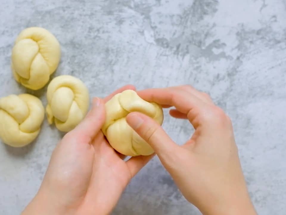 Hand closing a knot of dough to create a roll shape, three challah knots unbaked on countertop in background.