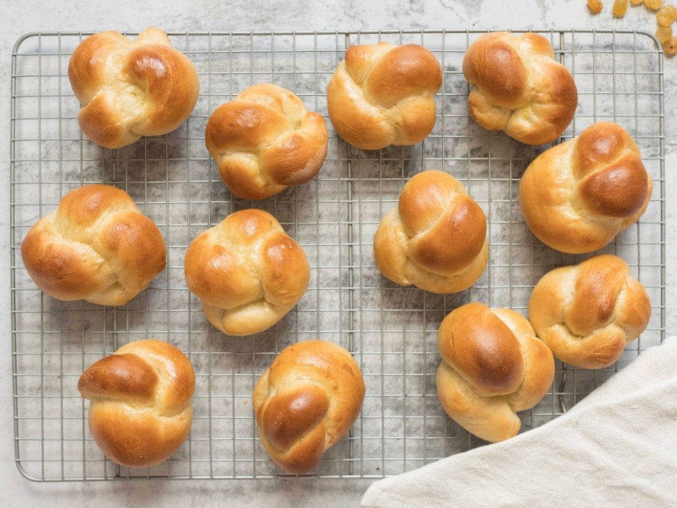 Golden brown challah rolls - knot-shaped baked rolls on wire cooling rack with linen cloth beside them.