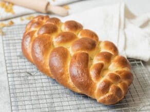 Horizontal shot of baked six strand challah on a wire cooling rack with linen napkin, rolling pin and raisins in background.