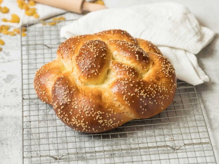 Horizontal shot of fully baked linked loops challah topped with sesame seeds, baked golden brown, on wire cooling rack with linen cloth, rolling pin and raisins in background.