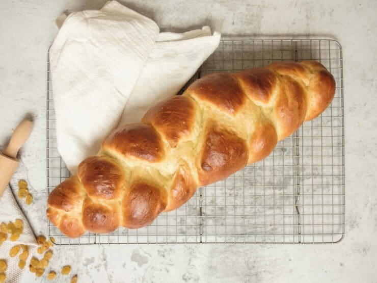 Overhead shot of fully baked 3 strand challah on cooling rack with linen cloth, raisins scattered on countertop.
