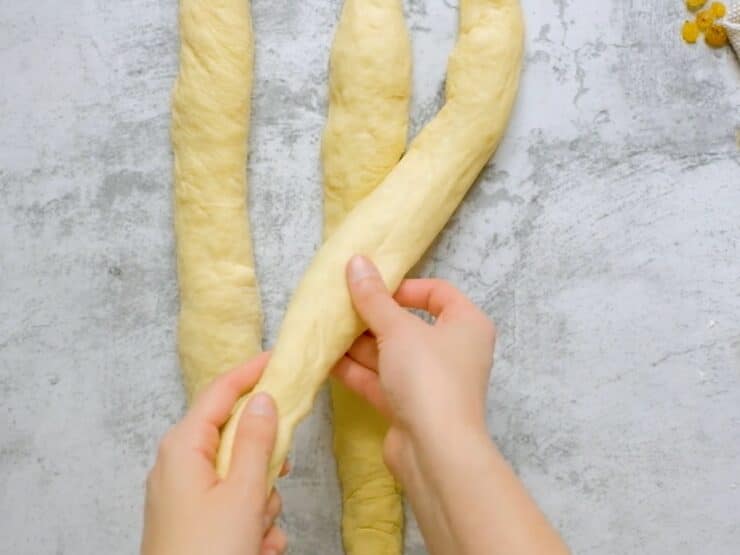 Overhead shot of three equal strands of challah laying next to each other. Two hands are crossing the strand on the right over the strand in the center.