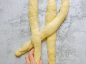 Overhead shot of three equal strands of challah laying next to each other. Two hands are crossing the strand on the right over the strand in the center and under the strand on the left.