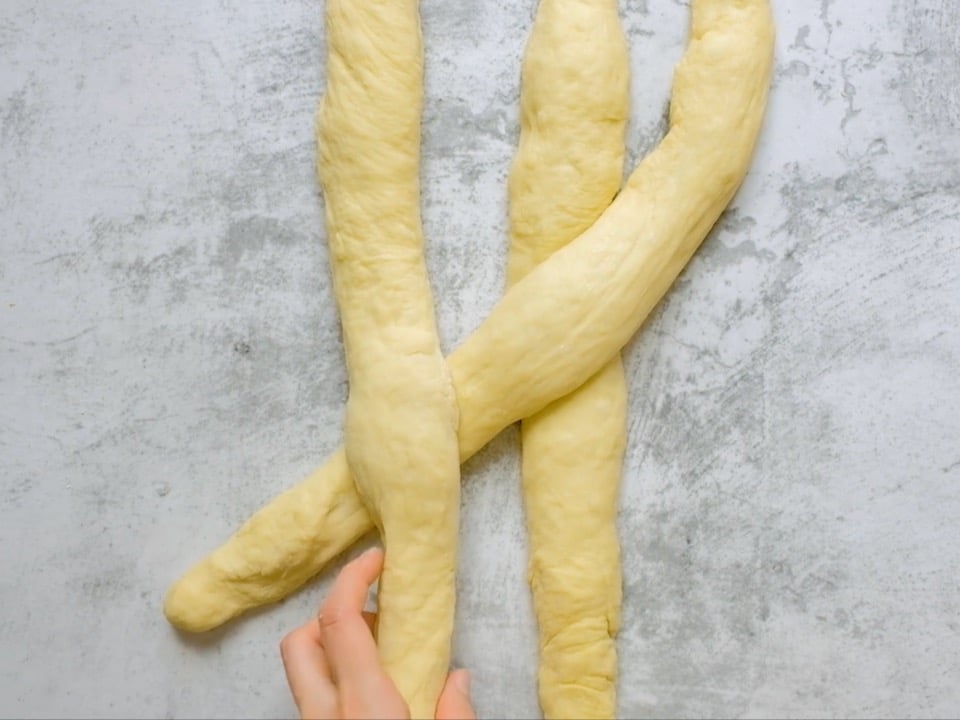 Overhead shot of three equal strands of challah laying next to each other. Two hands are crossing the strand on the right over the strand in the center and under the strand on the left.