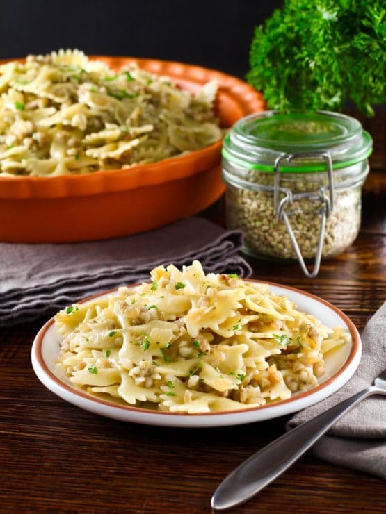 Vertical shot of Kasha Varnishkes with fresh parsley garnish. Larger serving dish, canister of kasha buckwheat groats, and fresh parsley sprig in background.