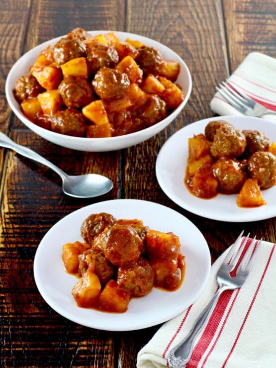 Plate of sweet and sour meatballs on wooden table with cloth napkin, large bowl of meatballs and spoon in background.