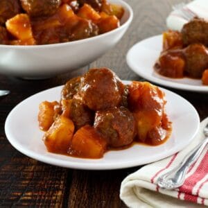 Sweet and sour meatballs on a plate with cloth napkin beside, more meatballs in dish in background.