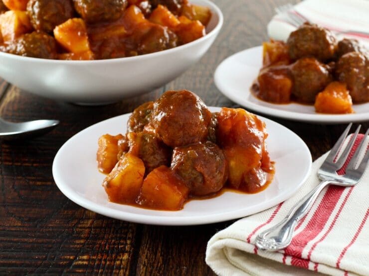 Sweet and sour meatballs on a plate with cloth napkin beside, more meatballs in dish in background.