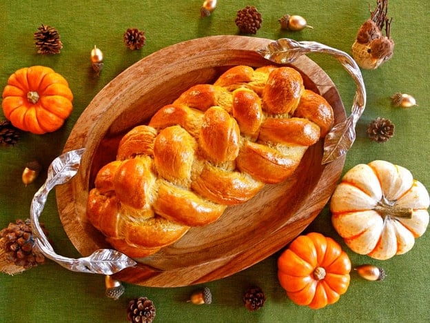 Golden baked Pumpkin Challah on top of a wooden platter with silver decorative handles on a green background. Small, decorative acorns, pine cones, and pumpkins are laying around the platter.