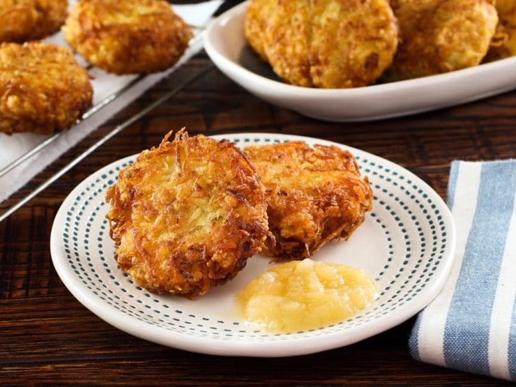 Plate with two crispy potato latkes and applesauce on table with cloth napkin, platter of latkes in background.