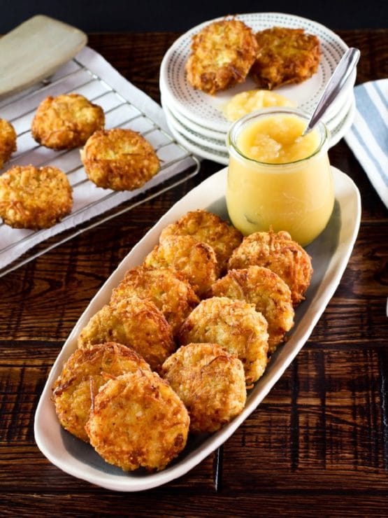 Vertical shot - long platter of classic potato latkes with carafe of applesauce and spoon on wooden table. Rack of latkes and plates stacked with a few latkes in background, along with cloth napkin.