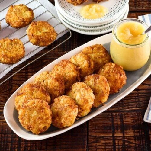 Horizontal shot - platter of classic potato latkes with carafe of applesauce and spoon on wooden table. Rack of latkes and plates stacked with a few latkes in background, along with cloth napkin.