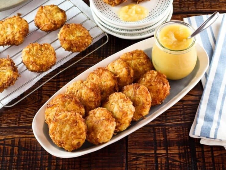 Horizontal shot - platter of classic potato latkes with carafe of applesauce and spoon on wooden table. Rack of latkes and plates stacked with a few latkes in background, along with cloth napkin.