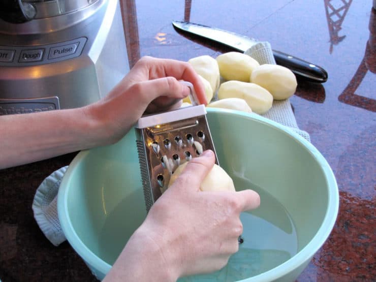 Hand grating peeled potatoes over an aqua-colored bowl next to a food processor, knife in background.