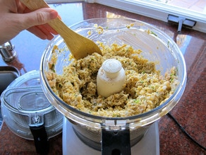 Falafel ingredients processed to a crumb-like texture in food processor, hand with wooden spoon scraping edges.
