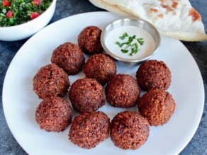 Horizontal shot - plate of several crisp, brown falafel balls with tahini sauce and fresh parsley garnish. In background, a large piece of laffa bread, and a bowl of tabouli salad.
