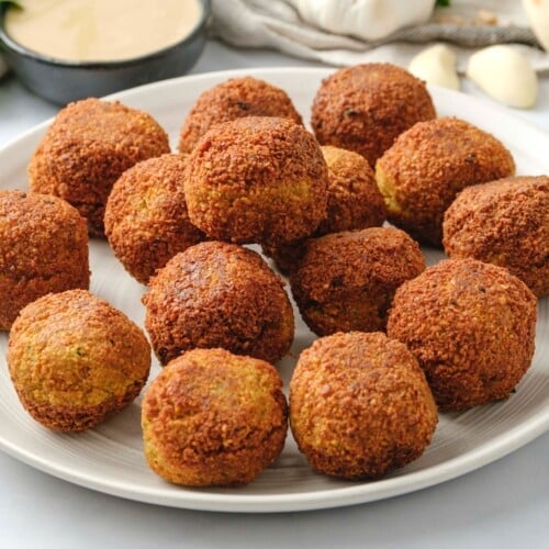 Horizontal image of a white plate filled with falafel balls. A bowl of tahini sauce, fresh herbs, a head of garlic, and pita bread sit in the background.