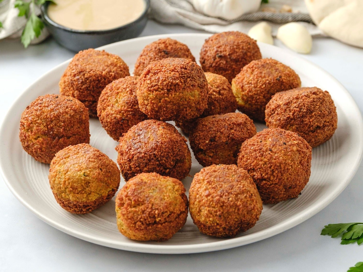 Horizontal image of a white plate filled with falafel balls. A bowl of tahini sauce, fresh herbs, a head of garlic, and pita bread sit in the background.