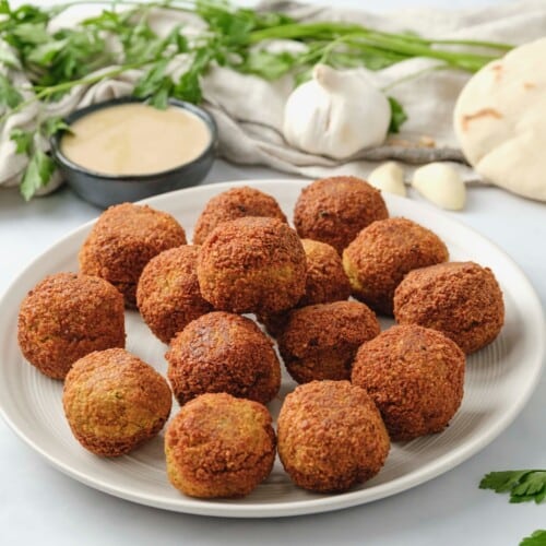 Horizontal image of a white plate filled with falafel balls. A bowl of tahini sauce, fresh herbs,a head of garlic, and pita bread sit in the background.