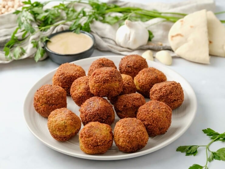 Horizontal image of a white plate filled with falafel balls. A bowl of tahini sauce, fresh herbs,a head of garlic, and pita bread sit in the background.