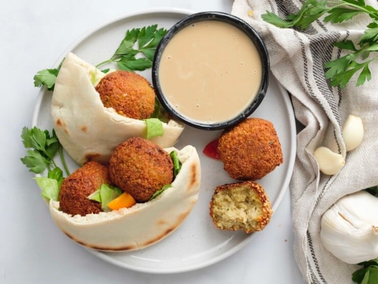 Horizontal overhead image of a plate containing a pita pocket filled with falafel next to a small black dish of tahini sauce.