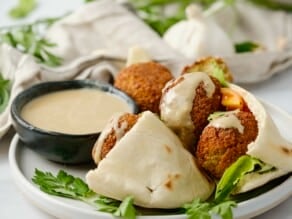 Horizontal close up image of a plate containing a pita pocket filled with falafel next to a small black dish of tahini sauce.