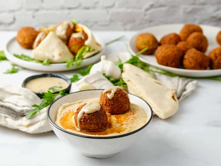 Horizontal image of a bowl of hummus topped with two falafel balls. A plate of falafel balls sits i the background next to a falafel pita sandwich.