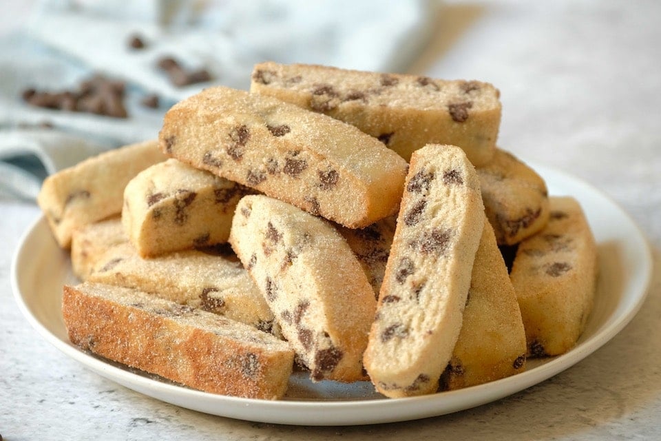 Horizontal shot - plate piled high with Jewish mandel bread cookies, aka mandelbrot, with towel and chocolate chips in background, on a marble countertop.