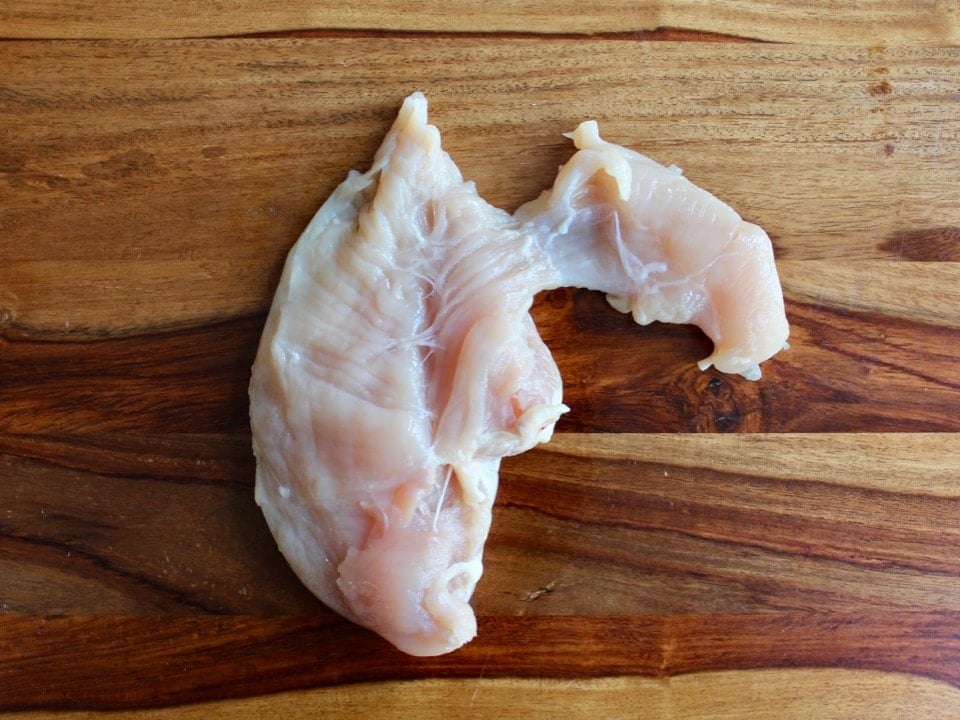 Chicken breast with tenderloin attached on wooden cutting board.
