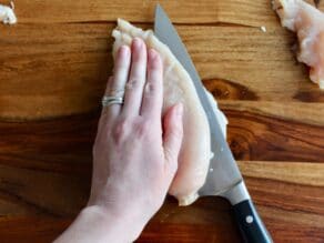 Chicken breast being sliced butterfly-style, hand holding down flat on top, on wooden cutting board.