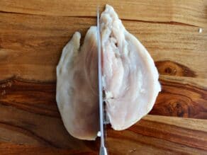 Butterflied chicken breast being sliced in half by chef's knife on wooden cutting board.