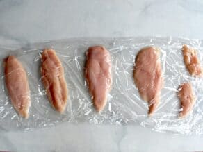 Four chicken breast halves and smaller tenderloin pieces between plastic wrap sheets on marble counter surface.