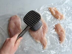 Mallet pounding chicken breast halves between two plastic sheets on marble countertop.