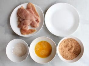 Five dishes - raw chicken breast halves, flour, beaten egg, breadcrumbs, and empty plate.