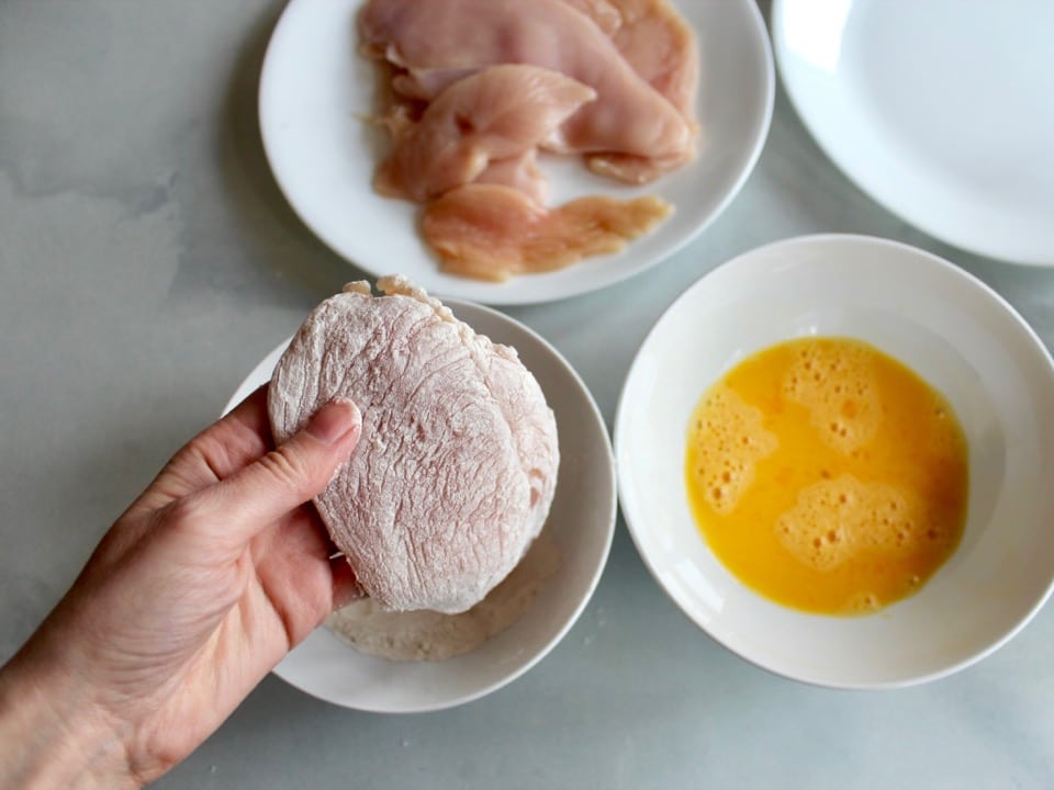 Chicken breast being dipped by hand into dish of flour. Dish of beaten egg and dish of raw chicken in background.