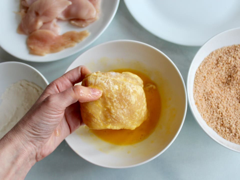 Flour dredged chicken breast being dipped by hand into dish of beaten egg.