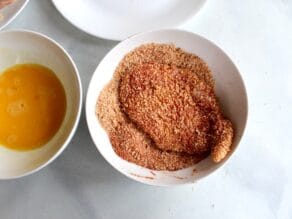Chicken breast resting in dish of paprika sesame breadcrumbs, fully breaded.