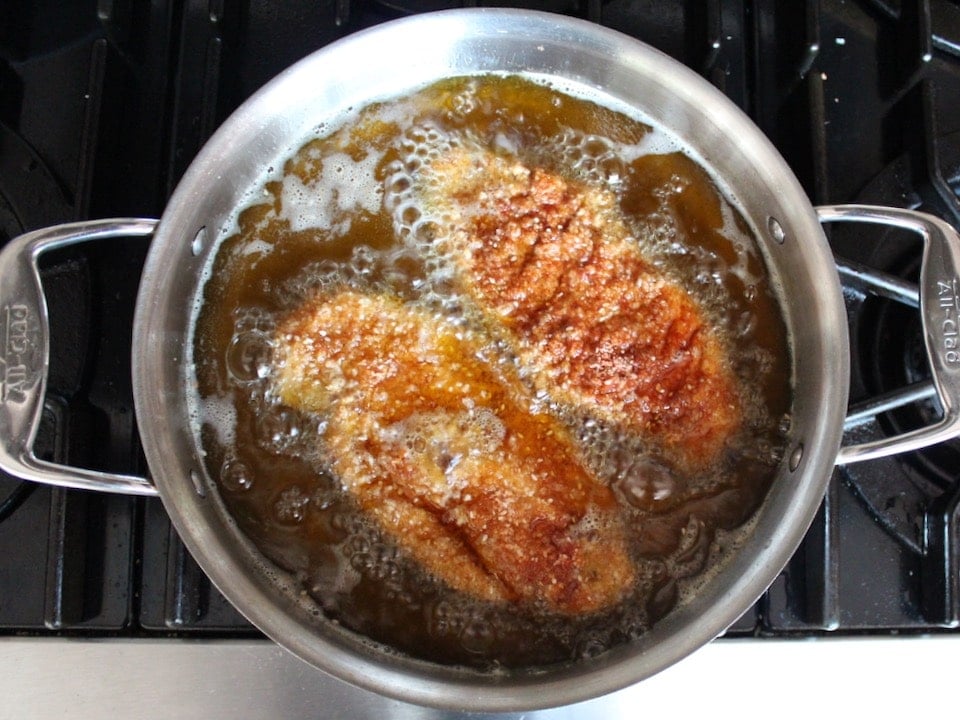 Two chicken schnitzels half cooked in a pan of bubbling oil, frying on stovetop.