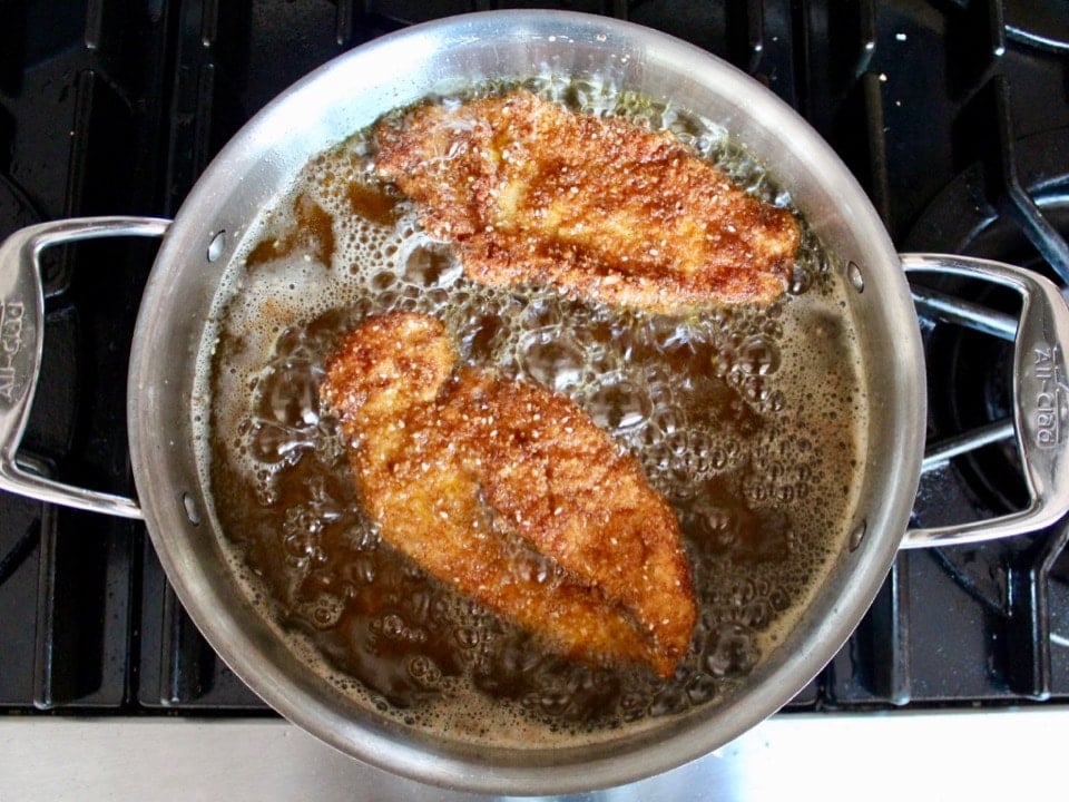 Two chicken schnitzels fully cooked in a pan of bubbling oil, frying on stovetop.